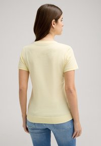 Gelbes Kurzarm-T-Shirt mit Rundhalsausschnitt und glatter Textur. Getragen mit hellblauen Jeans, Blick von hinten.