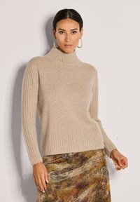 Beiger Rollkragenpullover mit geripptem Textur und langen Ärmeln, kombiniert mit einem rock mit metallischem Muster in erdigen Farbtönen und seidig schimmerndem Finish.