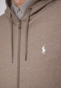 Felpa con zip marrone realizzata in tessuto morbido, dotata di una zip anteriore e cordini, con un piccolo logo bianco del polo sul petto.