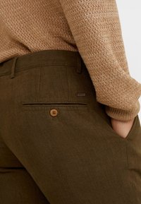 Olijf groene katoenen blend broek met een achterzak, houten knoopsluiting en subtiele textuur, gecombineerd met een lichtbeige gebreide trui.