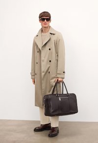 Beige, kétsoros trench kabát, kötött krém svédpulóver, krém nadrág, barna bőr cipők, fekete napszemüveg, sötétbarna bőr táska a kezében.