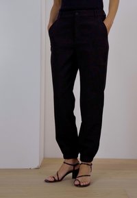 Black tailored trousers with a relaxed fit, featuring side pockets and a button closure, paired with strappy black heels on a wooden floor.