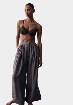 Mujer que lleva un sujetador negro y pantalones grises anchos de talle alto y sueltos, de pie con una mano en el bolsillo contra un fondo liso.