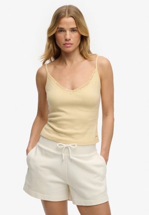 Kvinde med blondt hår iført en beige ribstrikket camisole med blonder og hvide shorts med snøre i taljen, hænderne i lommerne, neutral baggrund.