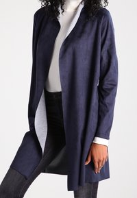 Marinblå lång jacka i mjukt mockaliknande material med en kontrasterande ljusgrå insida. Har öppen fram och avslappnad passform.