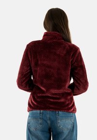 Giacca in felpa bordeaux con un motivo testurizzato, colletto alto e maniche lunghe, abbinata a jeans in denim azzurro chiaro.