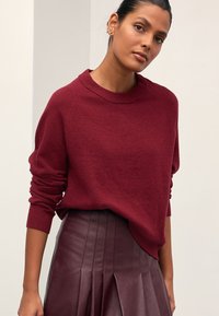 Bordeauxfarvet striktrøje med rund halsudskæring og afslappet pasform, kombineret med en bordeauxfarvet plisseret lædernederdel. Blød tekstur og ribstrikkede manchetter.