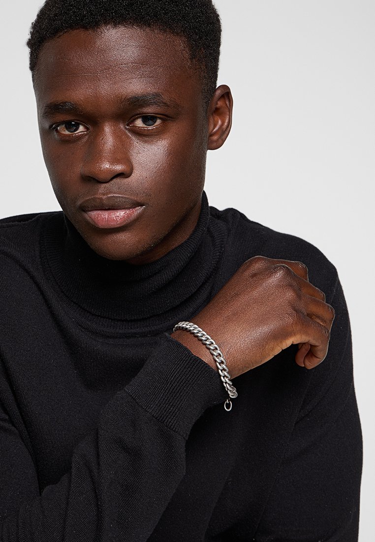 Joven en cuello de tortuga negro mostrando una pulsera de cadena de plata en la muñeca contra un fondo gris liso.