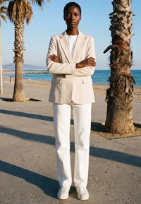 Femme en blazer beige, pantalon blanc et baskets, debout les bras croisés sur une promenade ensoleillée au bord de la plage entre des palmiers.