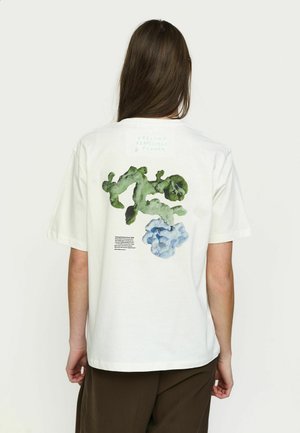 Person med langt brunt hår iført hvid T-shirt med grønt og blåt abstrakt blomsterdesign og lille trykt tekst på bagsiden.