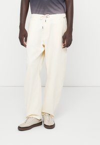 Pantalon en coton crème avec une coupe décontractée, doté d'une taille élastique avec cordon de serrage et de poches latérales. Associé à des baskets beiges.
