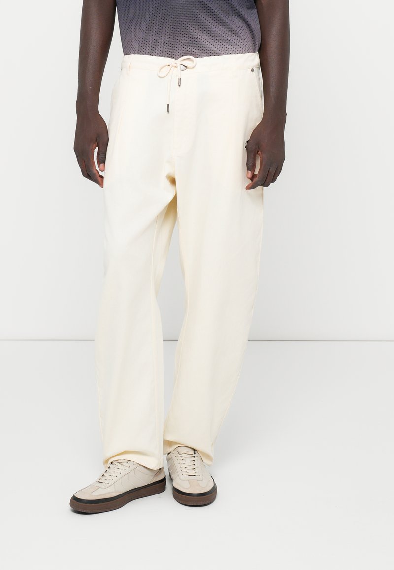 Pantalon en coton crème avec une coupe décontractée, doté d'une taille élastique avec cordon de serrage et de poches latérales. Associé à des baskets beiges.