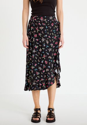 Czarna spódnica kopertowa z kolorowym, drobnym kwiatowym wzorem i falbanką u dołu, noszona z czarnym bezrękawnikiem i czarnymi sandałami z klamrą.