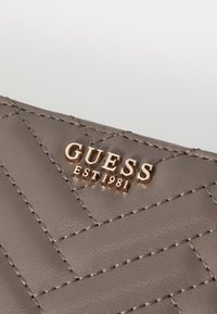 Billetera de cuero sintético gris con diseño acolchado, logo en relieve dorado "GUESS EST 1981" y detalles de costura en contraste.