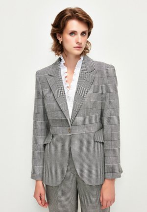 Blazer a rayas en gris y negro, con cintura ajustada, cierre de doble botonadura y cuello con muescas, combinado con una blusa blanca con volantes.