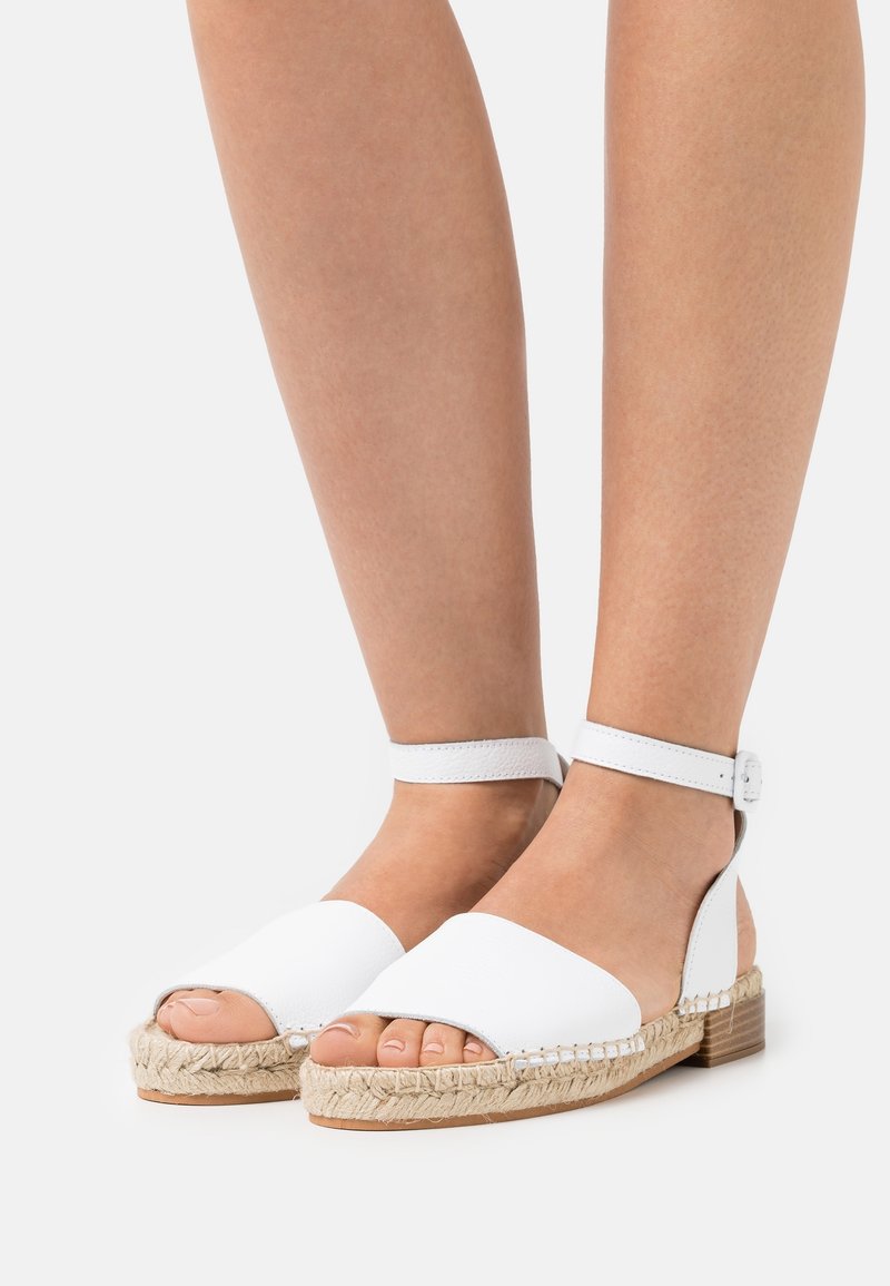 Pieds portant des sandales espadrilles blanches à bride à la cheville avec semelles en jute tressé et orteils découverts sur un fond uni.