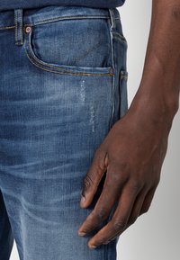 Jeans in denim di un blu medio con una texture liscia, caratterizzati da un classico design a cinque tasche e un leggero strappo sulla coscia.