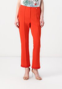 Rode op maat gemaakte broek met een soepele textuur, voorzien van een voorplooitje, een decoratieve knoop en gegolfde zoom, gecombineerd met naakte hoge hakken sandalen.