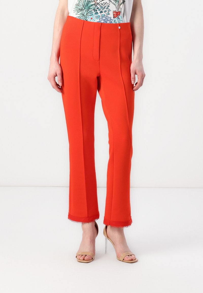 Rode op maat gemaakte broek met een soepele textuur, voorzien van een voorplooitje, een decoratieve knoop en gegolfde zoom, gecombineerd met naakte hoge hakken sandalen.