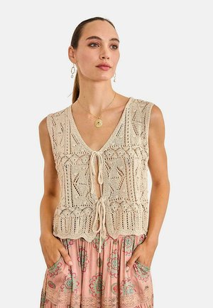 Mujer con chaleco beige de punto atado y falda rosa con estampado, lleva collar con colgante dorado y pendientes, manos en los bolsillos de la falda.