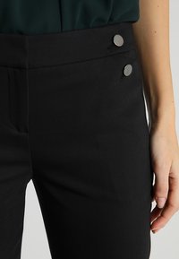 Pantalons noirs ajustés avec une texture lisse, dotés de deux boutons en métal argenté à la taille et d'une coupe cintrée.