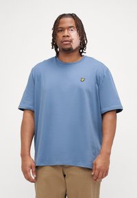 T-shirt en coton bleu à manches courtes et col rond, arborant un petit logo jaune sur la poitrine gauche. Coupe simple et décontractée.