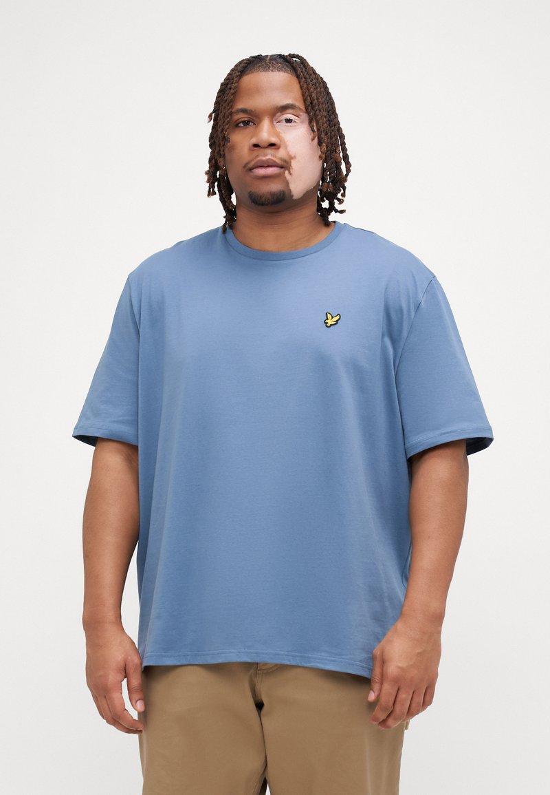 T-shirt en coton bleu à manches courtes et col rond, arborant un petit logo jaune sur la poitrine gauche. Coupe simple et décontractée.