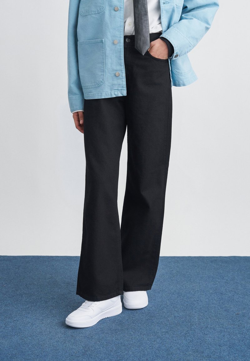 Persoon die zwarte wijde broek, witte sneakers, lichtblauwe jas, wit overhemd en donkere stropdas draagt, staand op een blauw tapijt.