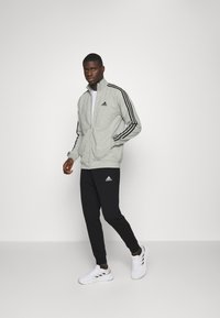 Siva zip-jakna z zadrgo z črnimi črtami na rokavih, v kombinaciji s črnimi hlačami in belimi supergami, na vsakem delu pa je vidna Adidasova oznaka.