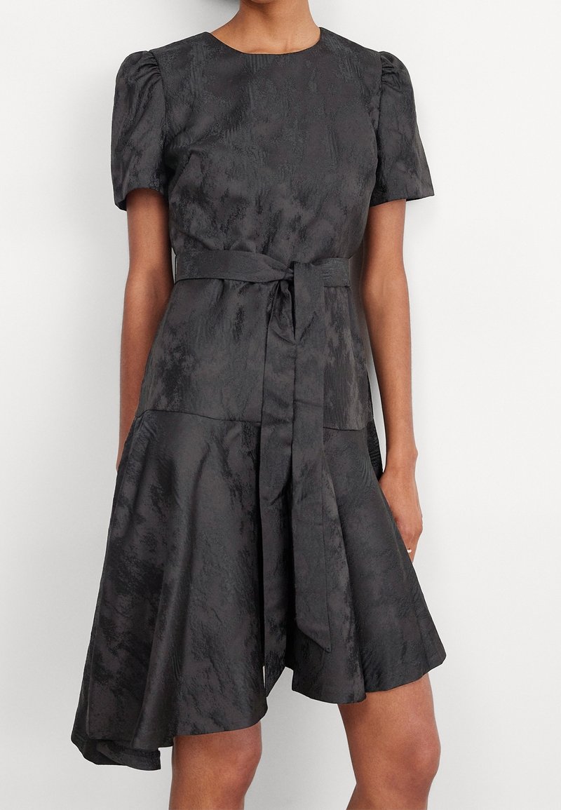 Robe gris foncé en tissu texturé. Manches courtes bouffantes, encolure ronde et ceinture à la taille. Jupe évasée avec un ourlet inégal.