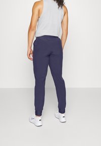 Person klädd i marinblå joggarbyxor, vita Nike-sneakers och en ljusgrå ärmlös toppen, stående mot en enkel vit bakgrund.