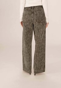 Personne portant un pantalon large à imprime léopard et un haut beige à manches longues, se tenant avec les mains détendues le long du corps devant un fond uni.