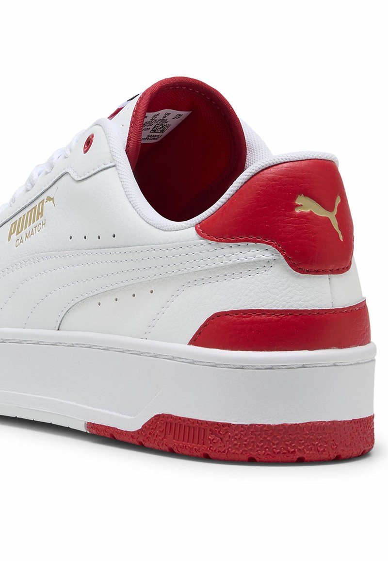 Baskets en cuir blanc avec des accents rouges sur le talon et la languette, présentant un logo doré, des détails perforés et une semelle extérieure texturée rouge.