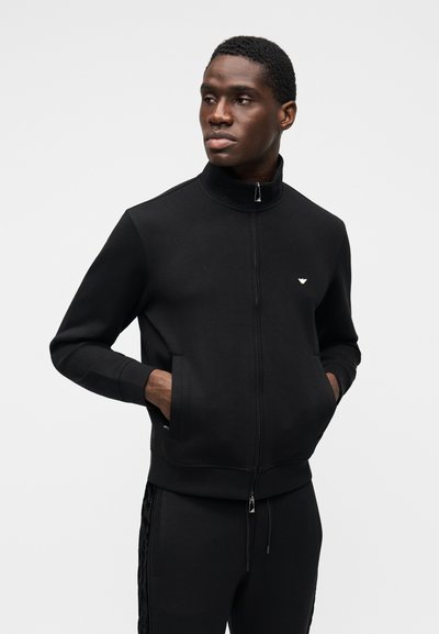 Juoda džinsinė striukė su aukštu apykakliu ir šoninėmis kišenėmis, pagaminta iš minkštos medžiagos. Yra subtilus logotipas ant krūtinės ir apsiūtas rankogalis.