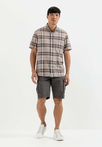Kariertes kurzärmliges Hemd in Grau, Orange und Weiß; kombiniert mit grauen Cargo-Shorts und weißen Sneakers. Auffällige Textilmuster und lässige Passform.