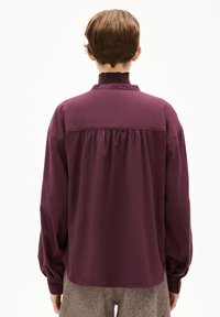 Top a maniche lunghe in un profondo bordeaux con una spalla arricciata e collo alto. Il tessuto sembra morbido con una vestibilità rilassata e maniche con polsini.