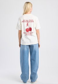 Cream-colored t-shirt features red cherry graphic and text. Paired with light blue, wide-legged jeans. Casual design, short sleeves.