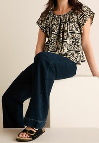Top a maniche corte nero e crema a fantasia con scollatura arricciata, abbinato a jeans a gamba larga blu scuro e sandali neri con fibbia dorata.