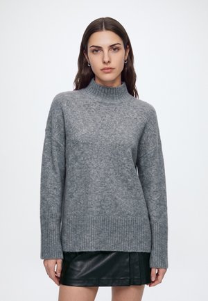 Femme aux longs cheveux bruns porte un pull à col roulé côtelé gris et une mini-jupe en cuir noire, debout devant un fond blanc uni.