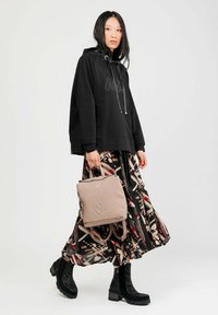 Mujer vestida con sudadera negra, falda midi estampada, botas negras hasta el tobillo, sosteniendo una mochila beige, de pie contra un fondo claro y liso.