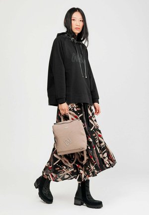 Frau in schwarzem Hoodie, gemustertem Midi-Rock, schwarzen Stiefeletten, die einen beigen Rucksack hält und vor einem schlichten hellen Hintergrund steht.