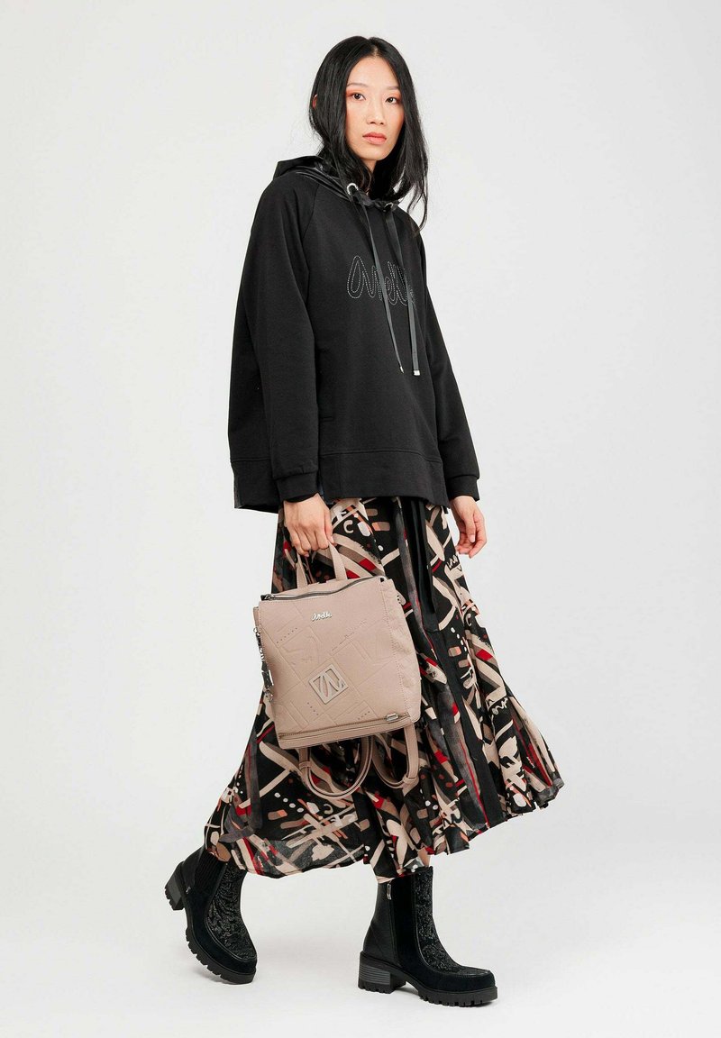 Mujer vestida con sudadera negra, falda midi estampada, botas negras hasta el tobillo, sosteniendo una mochila beige, de pie contra un fondo claro y liso.