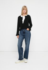 Frau steht mit einer Hand in der Tasche, trägt eine schwarze Knopfmanschette über einem weißen Hemd, blaue Jeans und weiße Sneakers, ein einfarbiger Hintergrund.