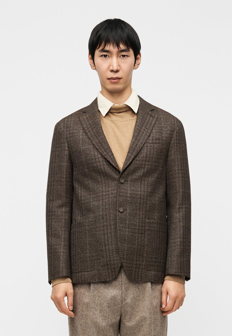 Καφέ καρό σακάκι με δύο κουμπιά, γιακάδες με εγκο πή και τσέπες με πατς, φορεμένο πάνω από μπεζ πουλόβερ και λευκό πουκάμισο με γιακά.