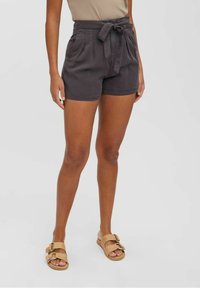 Personne portant un short taille haute gris foncé avec une ceinture nouée, un haut beige sans manches et des sandales beiges à double bride, debout sur un fond uni.