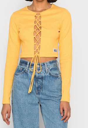 Top amarillo de manga larga y corte corto con detalle de cordones en la parte delantera, combinado con vaqueros de mezclilla azul claro de cintura alta.