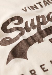 Kræmfarvet bomuldsstof med store, teksturerede mørkebrune bogstaver i vintage stil, der fremhæver ordet "Super".