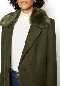Manteau vert olive avec col en fausse fourrure, design structuré, tissu texturé, fermeture à boutons sur le devant et poches latérales. Haut blanc en dessous.