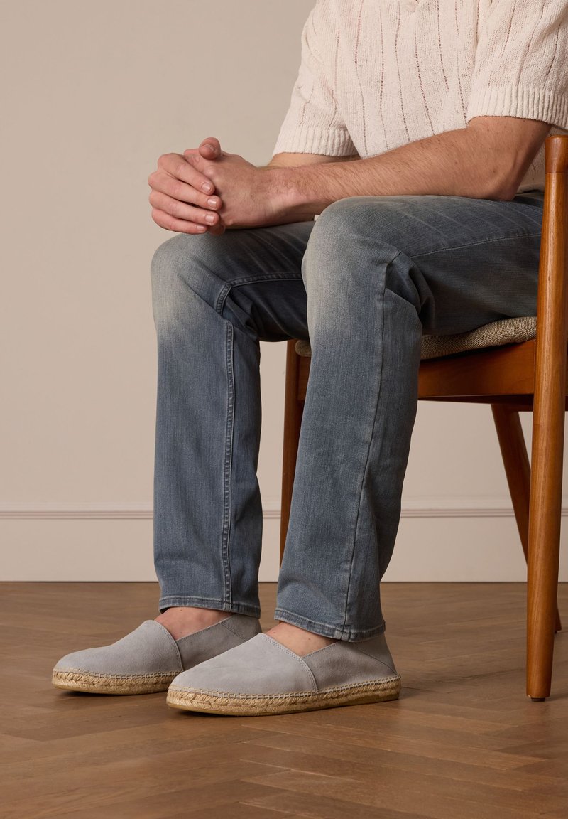 Espadrillas grigie con tomaia in suede e suola in giuta, indossate con jeans azzurri chiari, seduto su una sedia di legno.