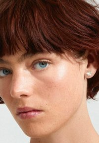 Silver stud earrings shaped like a butterfly with clear gemstone accents. The model has short, tousled red hair and light skin.